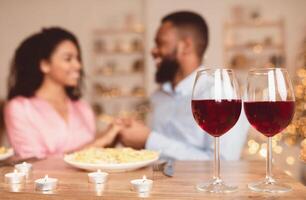 Romantic Date. Selective focus on two glasses of red wine on the table in the foreground. Black couple spending time together at home or cafe, having dinner, holding hands in blurred background photo