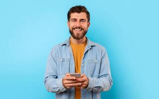 Cheerful guy with smartphone looking at camera and smiling, texting in social media over blue studio background. Positive young man in casual chatting on phone, using new mobile application photo