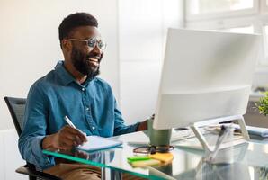 Positive black businessman writing notes in notebook while watching webinar course, looking at computer monitor and smiling, sitting at workplace in office photo