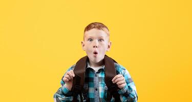 Amazed redhead schoolboy with backpack showing shocking face expression over yellow studio background, empty space. Cute ginger kid with eyes and mouth wide open looking at camera photo