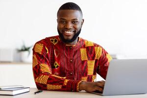 Portrait Of Smiling Black Freelancer Guy In African Traditional Clothes Sitting At Workplace In Home Office, Cheerful Man In Ethnic Costume Posing At Camera While Working With Laptop Computer photo