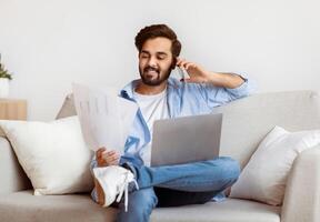 Eastern Freelancer Man With Cellphone, Papers And Laptop Working Remotely At Home. Millennial Arab Guy Reading Documents And Talking On Mobile Phone While Sitting On Couch With Computer, Free Space photo