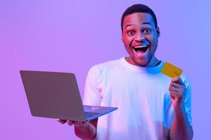 Online Shopping. Portrait of joyful black man with laptop and credit card standing in neon lighting over purple background, cheerful african guy enjoying making payments in internet, copy space photo