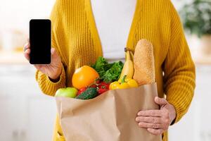 Cropped of woman with shopping bag full of grocery showing smartphone with empty screen, kitchen interior, copy space, mockup. Unrecognizable lady using mobile app for grocery shopping online photo