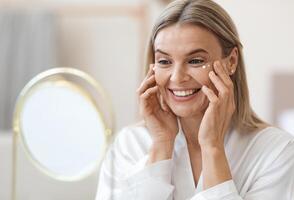 Happy beautiful blonde middle aged lady in silky bathrobe using moisturizing eye cream, looking at mirror and smiling, enjoying face care routine, closeup shot. Dark circles, eye wrinkles treatment photo