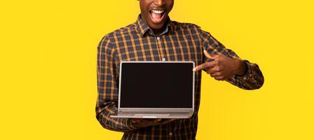 Check This Website. Joyful African Guy Pointing At Laptop Computer With Black Blank Screen, Standing Isolated Over Yellow Background, Demonstrating Free Place For Your Design, Mockup With Copy Space photo