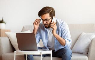 Arab Freelancer Man Talking On Cellphone And Working With Laptop At Home Office, Busy Eastern Guy Looking At Computer Screen While Discussing Project With Client, Sitting On Couch In Living Room photo