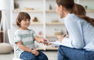 First meeting with patient. Friendly professional woman child psychologist greeting cute little boy, touching his hand and smiling at office photo
