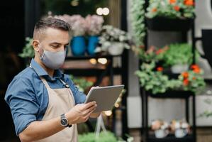 Small business, start-up, new normal and lockdown. Millennial man works in own flower shop in apron and face shield looks at tablet and accepts online order at studio, copy space, outdoor photo