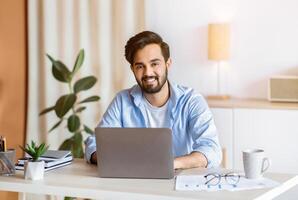 Remote Career. Happy Arab Male Entrepreneur Sitting At Desk With Laptop At Home Office, Looking And Smiling At Camera While Working Online On Computer, Eastern Man Enjoying Distant Job Opportunities photo