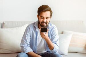 Smiling Arab Man Recording Voice Message On Smartphone Or Using Virtual Assistant While Relaxing On Couch At Home, Eastern Millennial Guy Enjoying Smart Technologies For Communication, Copy Space photo