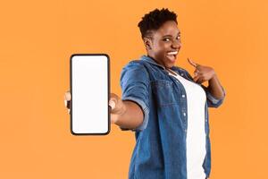 Mobile Application Ad. Excited African Woman Showing Phone Empty Screen Recommending App Standing Over Yellow Studio Background, Smiling To Camera. Cellphone Display Mockup, Check This Apps photo