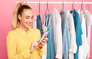 Buying Clothes Online Concept. Portrait of smiling lady holding and using mobile phone, shopping via internet. Influencer making purchases in virtual store, standing near hangers at pink studio photo