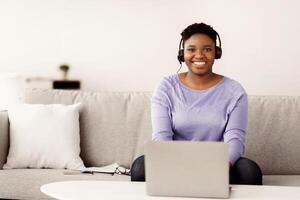 Portrait of smiling female customer service operator in headset posing at workplace, looking at camera, portrait of body positive hotline manager sitting on sofa with laptop in home office, copy space photo