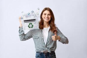 Waste Recycling Concept. Smiling young woman holding recycle bin with empty plastic bottles and showing thumb up gesture, eco-friendly lady standing over light background in studio, copy space photo