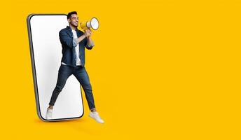 Handsome young arab man jumping out of huge smartphone with white empty screen, holding megaphone over yellow studio background, making important announcement, panorama with copy space photo