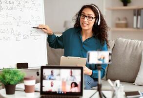 Videoconference And Training. Class of diverse students learning remotely online using web app. Female teacher in headset providing lesson looking at pc screen, recording on cellphone on tripod photo