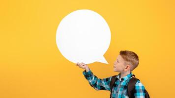 Side view of ginger boy schooler with backpack saying something loud and empty communication cloud over yellow studio background, copy space. Boy with schoolbag advertising something photo