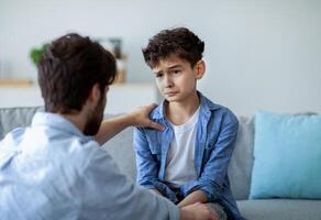 Father and sad son having heart to heart talk at home, daddy trying to solve problems, searching solution together. Parent showing care and love, calming down boy and holding hand on shoulder photo