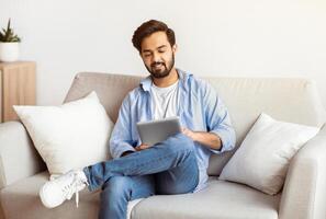 Modern Technologies For Leisure. Smiling Arab Man Using Digital Tablet At Home While Relaxing On Couch In Living Room, Eastern Millennial Guy Shopping Online Or Browsing Internet, Copy Space photo