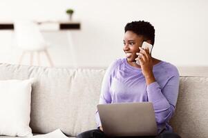 Making Business Call. Smiling african american woman talking on her mobile phone sitting on the sofa at living room with computer laptop, female working remotely, looking away at window photo