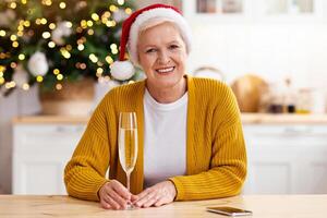 Monitor pov of smiling mature woman in Santa hat having online party with her family on New Year, sitting at kitchen, having xmas tree behind, holding glass of champagne, New Year during lockdown photo