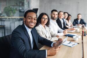 African American Businessman On Business Conference, Smiling To Camera During Corporate Meeting With Colleagues And Partners Sitting Together In Modern Office. Successful Business Career Concept photo