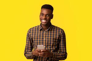 Portrait of happy young black guy with smartphone in hands smiling at camera, enjoying mobile communication and modern technologies while standing isolated over yellow studio background, copy space photo