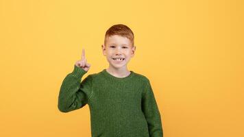 Happy ginger kid cute school boy in knitted green sweater pointing finger up over yellow studio background. Smiling redhead boy aiming up, having great idea or found creative solution photo