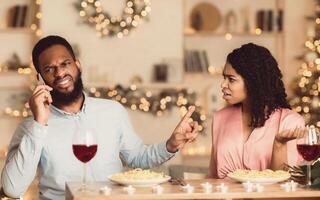 Annoyed african american woman looking at her man who talking on mobile phone and showing her finger to shut up and to wait while having date in restaurant, conflicts in couple concept photo