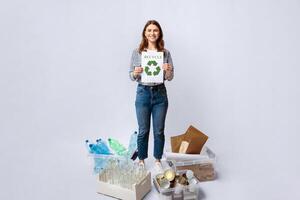 A woman stands confidently in a studio, holding a recycling sign while surrounded by various recyclable materials, including bottles, cardboard, and cans, showcasing her commitment to sustainability. photo