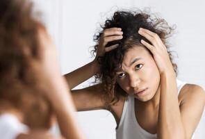 Afro-american girl frowning and looking intently on her face in mirror. Hair and sculp problems concept photo