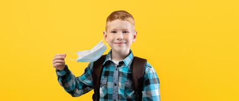 Smiling schoolboy taking off protective face mask over yellow studio background, copy space. Schoolboy holding face mask in hand, using protection against coronavirus while going to school photo