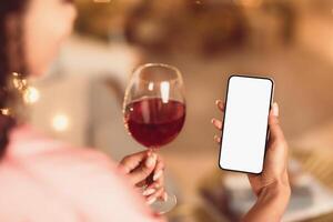 Back Over The Shoulder Back View Of Young Black Woman Holding And Using Mobile Phone With White Empty Screen For Mock Up, Making Virtual Call Or Watching , Drinking Red Wine From Glass photo