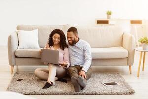 Virtual Communication. Happy black couple having online conference with friends or family using computer, waving hand to web camera, sitting on the floor carpet in bedroom at home photo