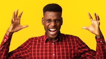 Portrait of young angry black man shouting with rage, furious african american guy raising hands and screaming with anger while standing isolated over yellow background, closeup with copy space photo