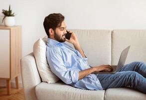 Freelance Concept. Handsome Arab Man Using Laptop And Talking On Cellphone At Home, Millennial Eastern Guy Working Remotely While Sitting On Couch In Living Room, Enjoyin Distant Job, Side View photo