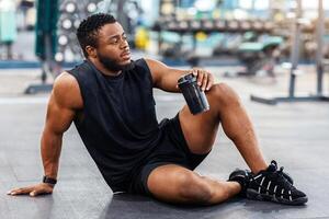 Handsome african american bodybuilder holding bottle with water or sports drink, sitting on floor at gym, resting, empty space. Smiling black muscular man having break while exercising at modern gym photo