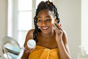 Daily beauty routine. Happy black lady applying moisturising cream on face, looking at magnifying mirror in bedroom, using nourishing skin and smiling, free space photo