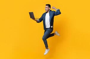 Emotional arab young man jumping up, using new modern laptop, raising fist up and smiling over yellow studio background, trading online, copy space for text or advertisement, panorama photo