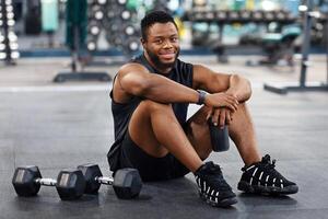 Smiling black muscular man having break while exercising with dumbbells at modern gym, copy space. Happy african bodybuilder holding bottle with water or sports drink, sitting on floor at gym, resting photo