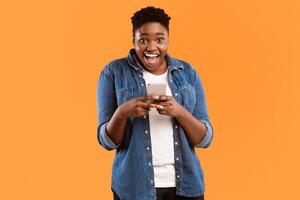 Great Application. Joyful Black Female Using New Smartphone And Mobile App Posing Smiling To Camera Standing Over Yellow Studio Background. Woman Playing Games On Phone Concept photo