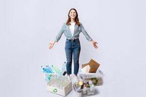 Waste Sorting. Happy woman standing among different kind of sorted garbage, showing boxes and containers with plastic, metal cans, glass bottles and paper for recycling over light studio background photo