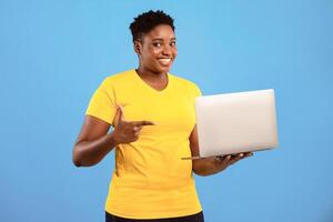 Oversized Black Woman Pointing Finger At Laptop Posing Holding Computer Advertising Website Standing In Studio Over Blue Background, Smiling To Camera. Online Service Ad photo