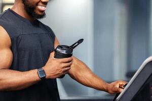 Cropped of muscular african american man having jogging work out, pressing on button. Unrecognizable black guy sportsman with protein drink standing on treadmill, side view, copy space, closeup photo