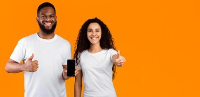 Smiling Multiethnic Couple Showing Cellphone Empty Screen Approving Application Gesturing Thumbs Up Standing Over Yellow Studio Background. We Like This Mobile App. Mockup, Panorama With Copy Space photo