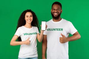 Charity Application. Two Diverse Volunteers Showing Smartphone Blank Screen Gesturing Thumbs-Up Standing Posing Over Green Studio Background. Great Volunteering App Concept. Mockup photo