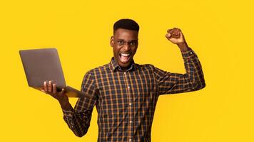 Euphoric Black Gguy Celebrating Success With Laptop, Raising Clenched Fist And Exclaiming With Excitement, Joyful African American Man Holding Computer, Standing On Yellow Background, Copy Space photo