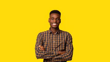 Positive Young Black Guy Posing With Folded Arms Over Yellow Studio Backgound, Handsome Millennial African American Man Wearing Checkered Shirt, Smiling And Looking At Camera, Panorama With Copy Space photo