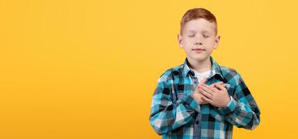Adorable redhead boy with eyes closed holding both hands on chest over yellow studio background, praying or feeling thankful, panorama with empty space. Peaceful kid with palms on heart photo